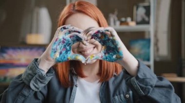 Ön manzara, 20 'li yaşlardaki kızıl saçlı, gözlüklü, gülümseyen, dişlek, aşk ressamı kadınla gülümseyen, elinde boyalı ellerle kameraya bakan, kalp şeklinde nazik bir jest yapan bir kız.