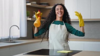 Neşeli kadın hizmetçi güzel anne Arap temizlikçi önlük giyip eldiven takıyor modern apartman içinde hijyenik sprey sıkarak temiz mutfağa taşınıyor müzik zevkli ev işleri yapıyor.
