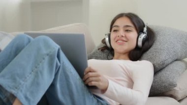 Kaygısız, gevşemiş Arap kız, kulaklıklı serbest çalışan öğrenci kanepe başında dinleniyor, kamera önünde konuşuyor, şarkı söylüyor, evden eğitim amaçlı internet üzerinden sohbet ediyor.