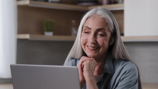 陽気な幸せな高齢女性ビジネス女性の祖母で無線イヤフォントークでコンピュータウェブカメラ笑みを浮かべて孫とチャットリモートでオーディオレッスンを聞くビデオをオンラインでホームキッチン — ストック動画