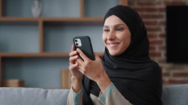 Siyah tesettürlü, rahat Müslüman bir kadın evinde akıllı telefon kullanıyor, cep telefonuyla sosyal medyada geziniyor mobil uygulamalarla haberleri kontrol ediyor, cep telefonu teknolojisiyle yemek siparişini kontrol ediyor.