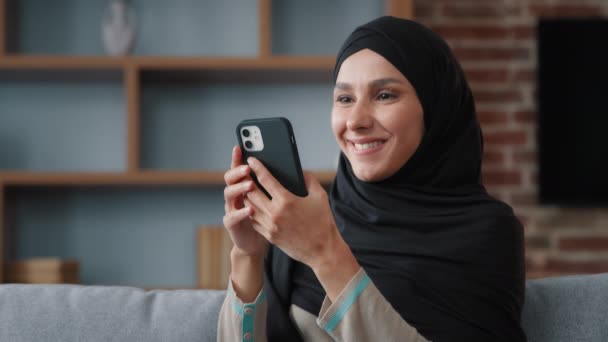 モバイルアプリでソーシャルメディアをサーフィン笑顔で自宅でスマートフォンを使用して黒ヒジャーブでリラックスしたイスラム教徒のイスラム教徒の女性は ニュースをチェックします携帯電話のガジェット技術サービスで食品配達注文を購入 — ストック動画