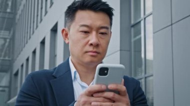 Alttaki görüntüde Asyalı yetişkin adam orta yaşlı 40 'lı yaşlarda işadamı ofis binasının yakınında dikiliyor. İnternetten akıllı telefon web sitesinden yazı yazarak sohbet etmeyi düşünüyor.