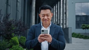Resmi takım elbiseli 40 'lı yaşlarda Asyalı yetişkin bir işadamı şehir merkezindeki akıllı telefona bakarak ofise gidiyor. Patron gülümsüyor ve cep telefonu ağını internette geziyor.