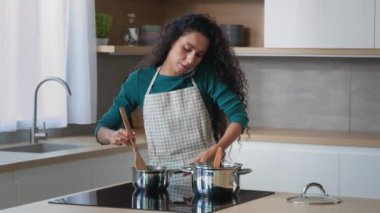 Meşgul Arap anne çekici kıvırcık ev hanımı şef evde kahvaltı hazırlarken önlük giyer tencerelerde yemek pişirir kaşıkla aile içi cep telefonu konuşmaları için yemek hazırlar.