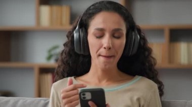 30 'lu yılların Latin kökenli beyaz kadın kulaklık takıyor. Evde eğleniyor, telefonuyla müzik dinliyor. Mobil uygulamayı ses için kullanıyor.