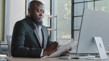 Kağıt tablet üzerine notlar yazan Afrikalı yazar ofis masasına oturur yetişkin girişimci Amerikalı iş adamı yeni fikirler yaratır şirket stratejileri pazarlama planı kontrol listesindeki noktaları doldurur