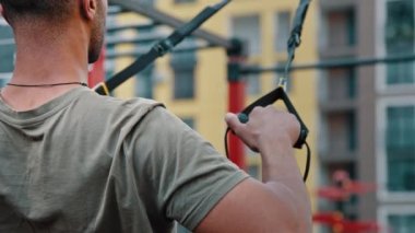 Asılan kaslı sporcu spor sahasında egzersiz yapıyor. Kol kaslarını çalıştırmak için spor kemerleri kullanıyor. Göğüs kaslarını çalıştırmak için emniyet kemerlerini kullanıyor.