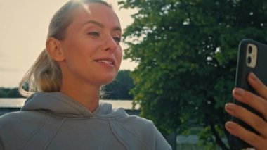 Gri kapüşonlu spor giysili formalı sporcu kadın sabah sporu eğitimi sırasında akıllı telefondan sokak ortasında sanal görüntülü konuşma yapıyor. Beyaz kadın sporcu hoş sohbetler yapıyor.