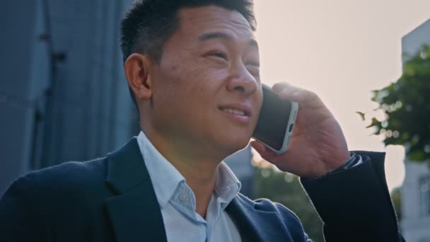側のビューを閉じる笑顔幸せなアジアの中高年実業家韓国の起業家の雇用主の男性フレンドリーなスマートフォンを呼び出すことによって話をする快適な携帯電話との会話を日光の下で — ストック動画