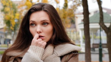 Doubtful pensive young woman brunette caucasian girl stand alone in autumn urban park holds hand to chin looking around waiting for boyfriend late feels lonely worries think difficult decision unsure