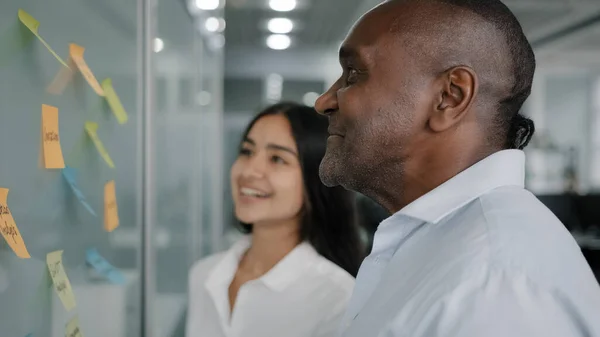 Corporate team business partners colleagues look at paper memo stickers on wall discuss project giving business advice managing tasks structure sticky notes brainstorming teamwork at office meeting