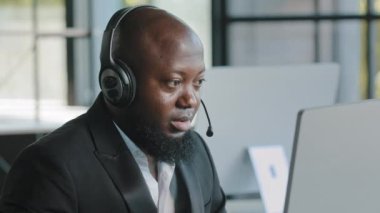 Yetişkin yönetici pazarlama uzmanı kulaklık takıp internetteki uygulamaları kullanarak internetteki odaklanmış Afrikalı Amerikalı çalışan ağlarda sohbet ediyor.