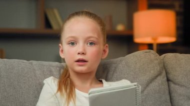 Genç kız anaokulu öğrencisi kanepede oturup not defterinde notlar okuyarak online edebiyat dersi ödevi hazırlıyor zeki okul çocuğu ev ödevini yalnız başına yapıyor webcam görüntüsü yapıyor.