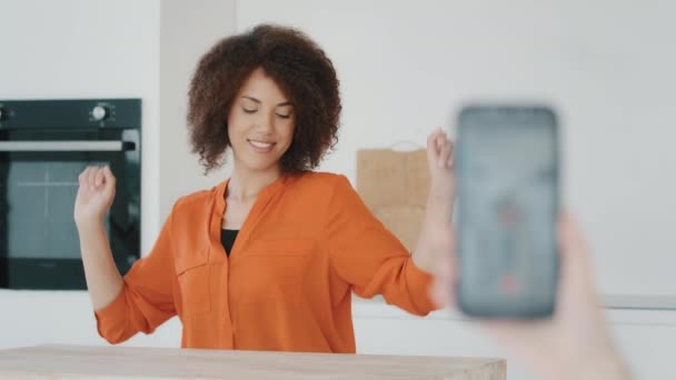 难以辨认的女摄影师手拿着手机在网上拍摄视频直播视频 与非洲卷曲女孩在厨房博客博客女性中跳舞 — 图库视频影像