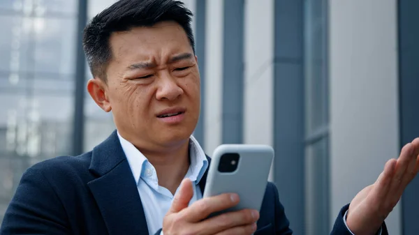 Close up Asian middle-aged adult man mad unhappy businessman feeling annoyed with using broken smart phone low battery problem angry having problem with mobile telephone frustrated bad news outdoors