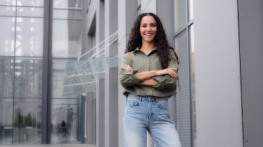 In city downtown background standing single smiling happy proud 30s woman in casual blouse strong confident pose with arms crossed female look at camera. Independent businesswoman portrait outdoors