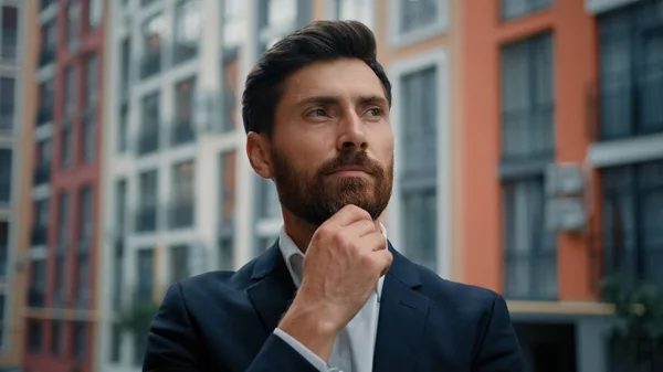 Caucasian smart puzzled thoughtful man hold hand to chin dreaming about career growth successful employer in stylish black suit stand outside modern office pondering idea planning business development