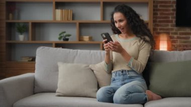 30 'lu yılların Latin Latin kökenli Kafkas kadın kanepede oturmuş cep telefonuyla oyun oynuyor. İnternet üzerinden zafer kazanmanın sanal hop e-ticaretinde indirimi var. Zaferi kutlayın.