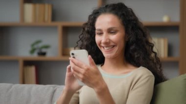 Gülümseyen, dişlek, beyaz kadın, kıvırcık saçlı, komik videolu, cep telefonlu, cep telefonlu, neşeli mobil uygulamaları izleyen Latin kadın.