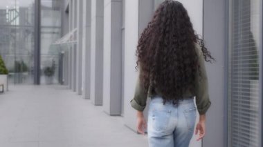 Back view from behind female with dark health curly long hair brunette woman businesswoman walker walking going in city near downtown company building turning head happy smiling looking at camera