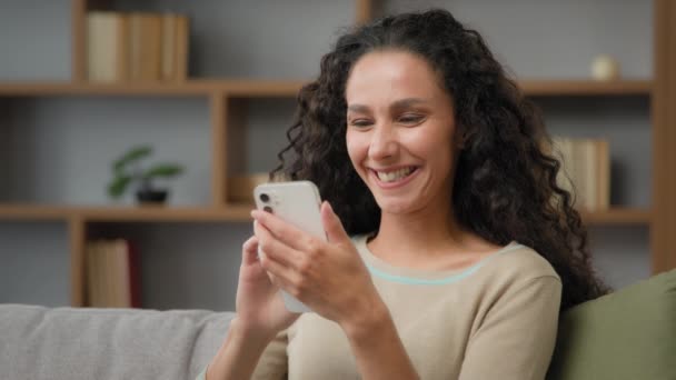 快乐的笑着笑着的白种人拉美裔女性卷发西班牙裔女性带着滑稽的视频在智能手机里拿着手机在家里沙发上看手机有趣的幽默应用程序 — 图库视频影像