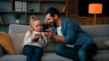 Beyaz bir adam ve küçük bir kız birlikte oturuyorlar modern telefonları dinliyorlar medya içeriklerine göz atıyorlar oyun oynuyorlar yetişkin baba çocuk kızına kablosuz cihazda komik videolar gösteren mobil uygulamalar kullanıyorlar.