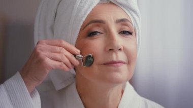 60 'lı yaşlarda, kafası havluyla banyoda yaşlı beyaz bir kadın taş patenciyle yüz masajı yapıyor. Güzel bayan cilt yogası prosedürü. Cilt bakımı için yüz bakımı.