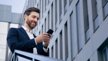 Elinde e-posta tutan şık yakışıklı işadamı sokakta gülümsüyor sosyal ağda mesaj iletileri yazıyor düşünceli adam akıllı telefondaki cep telefonu uygulamasını kullanarak online mağazada malları karıştırıyor.