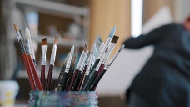 画笔工具画笔放在画室的桌子上 背景模糊了红头发的女画家在画布上画画 忙着创作爱好 — 图库视频影像