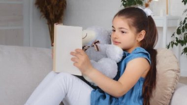 Kafkas Hindistanlı, İspanyol, çocuk, kız, öğrenci, oyuncak ayı, kanepeye oturmuş kitap okuyor, okul için evde eğitim ödevi okuyor.