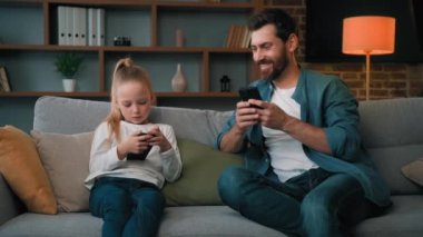 Beyaz bir adam ve küçük bir kız birlikte oturuyorlar modern telefonları dinliyorlar medya içeriklerine göz atıyorlar oyun oynuyorlar yetişkin baba çocuk kızına kablosuz cihazda komik videolar gösteren mobil uygulamalar kullanıyorlar.
