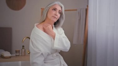 Banyoda bornozlu yaşlı beyaz kadın omuzlarına ve boynuna losyon sürmüş vitamin vitaminleri kolajeni yumuşak cilt yenilenmesi kırışıklık önleyici nemlendirici prosedür için yağ kullanıyor.