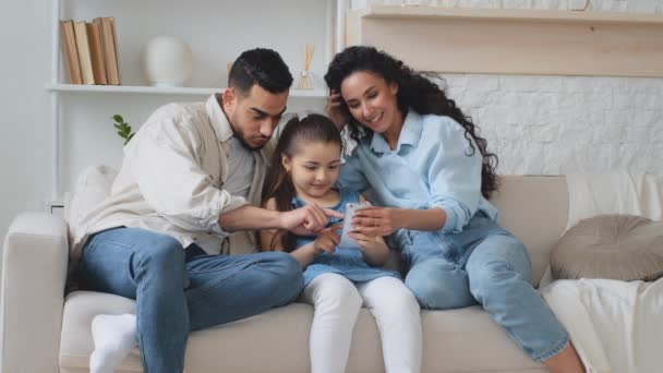 西班牙家庭坐在沙发上 用电话拍照和录像 父母带着小女孩玩的开心 试图用滑稽面具在网上移动应用程序笑孩子露出舌头 — 图库视频影像