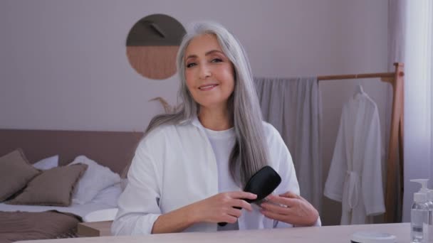 ポートレート高齢者熟女50歳白人女性でバスローブ朝の美しさルーチン見てカメラを持っています灰色の長い光沢のある髪と櫛の毛ブラシ健康的な髪型をお楽しみください — ストック動画