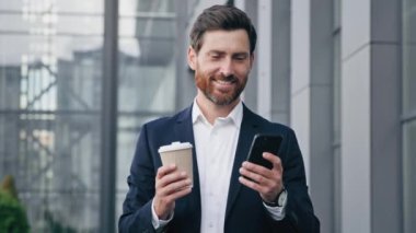 Başarılı gülümseyen yakışıklı işadamı elinde kahve bardağıyla sokakta yürüyor akıllı telefon ekranına bakıyor sosyal ağlara bakıyor cep telefonuyla internet üzerinden iyi haberler okuyor.