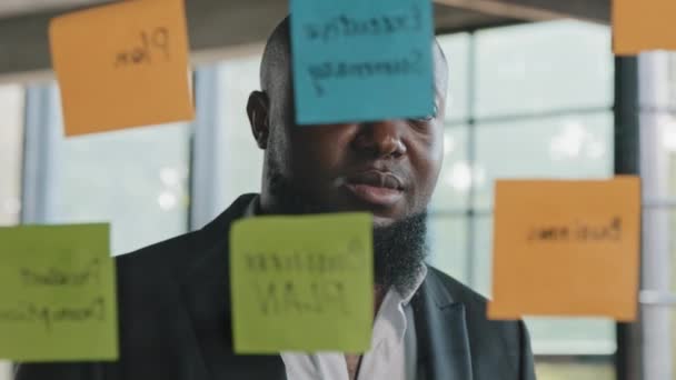 アフリカのビジネスマンのコーチの先生はオフィスでステッカーを読むガラスの壁にリマインダーを入れて創造的なビジネスアイデアを書く準備をする会社の戦略チェックの情報プロジェクトのタスクを粘着ノートにチェック — ストック動画