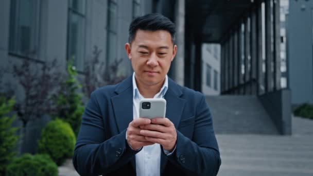 亚洲成熟的商人拿着智能手机站在城中笑着 中年经理首席执行官使用手机移动应用程序 工作时遥望着镜头微笑 — 图库视频影像