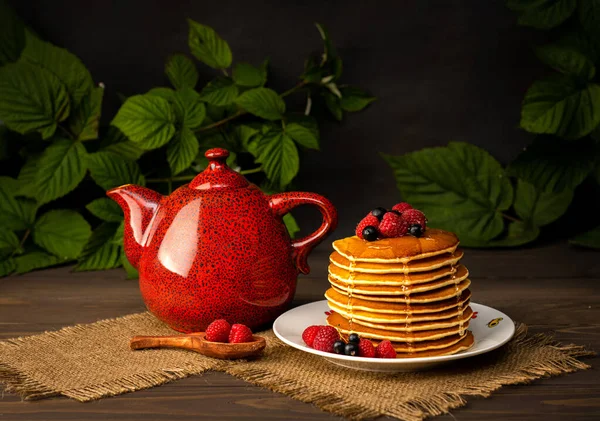 Pancakes desert with raspberries, blackcurrant and honey on white plate near red teapot on a black background. Healthy pancakes breakfast with berries on old wooden brown table.