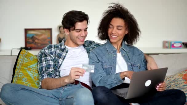 快乐的年轻的混血儿，和女孩坐在客厅的沙发上，穿着休闲时髦的衣服，用笔记本电脑和信用卡在网上购物，买东西，微笑 — 图库视频影像
