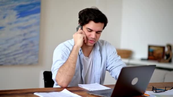 Homme latino intelligent positif, gestionnaire, designer ou pigiste vêtu de vêtements élégants décontractés, travaillant à la maison avec un ordinateur portable, parlant par téléphone portable avec un collègue, regardant à travers un projet d'entreprise — Video