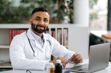 Karizmatik sakallı bir Hintli genel terapistin portresi, tıbbi üniforma ve steteskop içinde, kabinede, hastanede masasında oturuyor, kameraya bakıyor, dostça gülümsüyor.