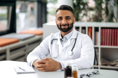 Yakışıklı bir Hintli genel terapistin portresi, tıbbi üniforma ve steteskop ile, masasında oturuyor, hastanede, kameraya bakıyor, dostça gülümsüyor.