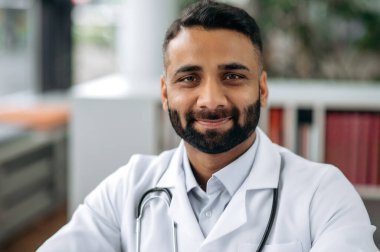 Tıbbi üniformalı ve steteskoplu bir Hintli genel terapistin yakın plan portresi. Kabinede, hastanede masasında oturuyor, kameraya bakıyor, dostça gülümsüyor.