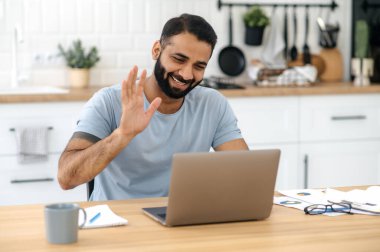 Çevrimiçi video bağlantısı. Pozitif başarılı Hintli adam, serbest çalışan, tasarımcı ya da yönetici, evden bilgisayar kullanarak iletişim kuruyor, iş arkadaşı ya da arkadaşıyla konuşuyor, gülümsüyor.