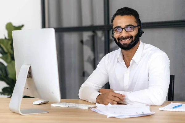 Retrato de un guapo empresario, gerente o especialista en TI indio inteligente, con una camisa blanca, con auriculares, sentado en su escritorio en la oficina, mirando a la cámara con una sonrisa amistosa — Foto de Stock