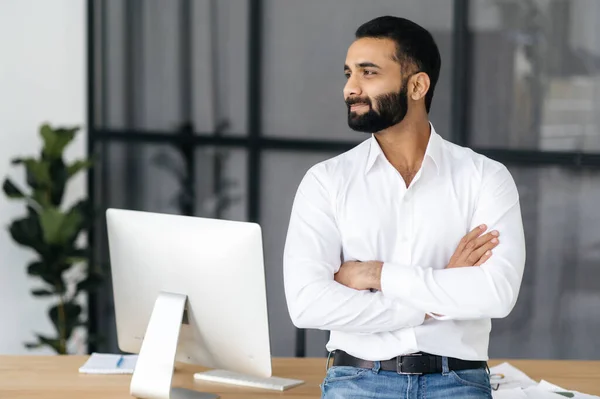 Retrato de um empresário indiano elegante confiante, gerente ou especialista em TI, vestindo uma camisa branca, de pé perto de sua mesa no escritório com os braços cruzados, olhando para longe e sorrindo — Fotografia de Stock