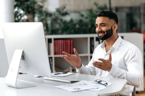 Amigável confiante bem sucedido indiano homem, homem de negócios ou mentor, sentado em sua mesa, vestindo uma camisa branca, usando computador para comunicação distante, conversando com um colega via videochamada, sorrindo — Fotografia de Stock