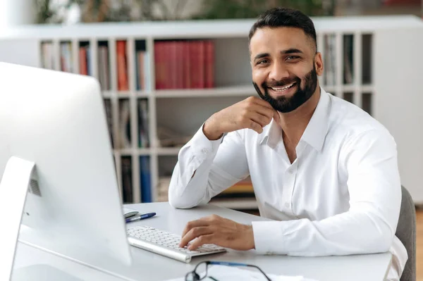 Retrato bonito empresário indiano, freelancer ou gerente, vestindo camisa branca, sentado à mesa com computador no escritório, olhando para a câmera e sorrindo amigável — Fotografia de Stock