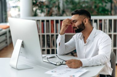 Yorgun Hintli bir adam, şirket sahibi ya da ofis çalışanı, çalışma masasında gözleri kapalı oturur, gözlerini ovar, baş ağrısı, stres ve yorgunluk hisseder, fazla mesaiden sonra dinlenmeye ve dinlenmeye ihtiyacı vardır.